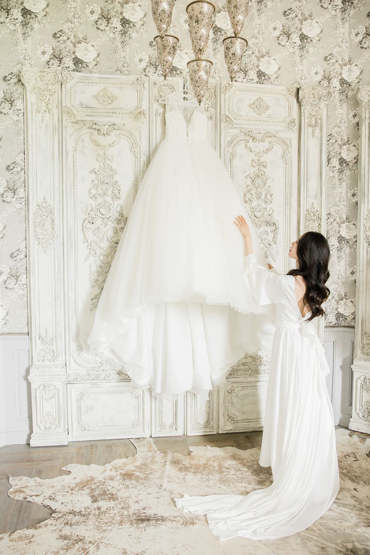 A getting-ready scene shot indoors in a ornate room featuring grey and white floral wallpaper, heavily carved white-painted panelling, and a gilded chandelier with warm bulb lighting. The bride, wearing a long white satin robe with wide sleeves and a bow tie at the waist, kneels on a cowhide rug in ivory and brown tones while reaching up to touch her wedding gown. The wedding dress is a voluminous ivory ballgown with a lace and beaded bodice and a full tulle skirt, hung from the chandelier. The overall decor palette is ivory, white, and soft grey, giving the room a classic French interior style. The shot is a medium-wide portrait framing both the bride and the full length of the hanging gown.