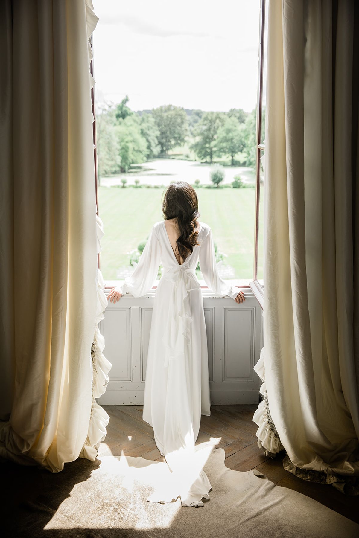 A bride stands with her back to the camera at a large open French window inside what appears to be a château, looking out over formal grounds with a lawn and mature trees in the distance. She wears a white long-sleeve wedding gown with a deep V-back, a bow tie detail at the waist, and a flowing train that pools on the herringbone parquet floor. Her dark hair is styled in a loose half-up wave. The room features floor-to-ceiling cream linen curtains framing the window, and natural light casts a sharp rectangle across the wooden floor. The composition is a full-length portrait shot from behind, centered symmetrically between the two curtain panels.