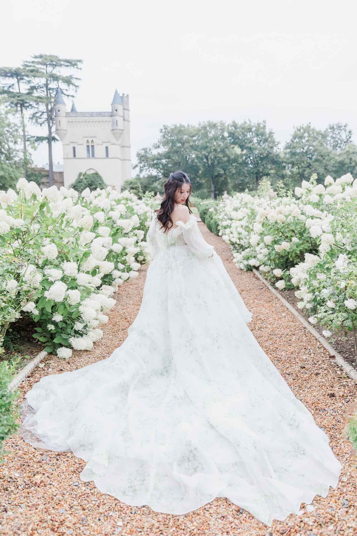 A bridal portrait shot outdoors in a formal château garden, with the bride positioned on a gravel pathway flanked by rows of white hydrangea bushes in full bloom. The bride wears an ivory off-the-shoulder ballgown with long sheer bishop sleeves, a floral-embroidered tulle skirt, and an exceptionally long cathedral train that extends the full length of the path toward the camera. She is photographed from behind, glancing over her shoulder with her dark hair styled in a loose half-up arrangement. A turreted château tower with Gothic arched detailing is visible in the background, slightly out of focus. The composition is a wide portrait shot taken from ground level along the central axis of the garden path, emphasizing the length of the train and the symmetry of the hydrangea borders. The overall styling is classic and romantic, with a soft, bright edit. Potential venue feature image.