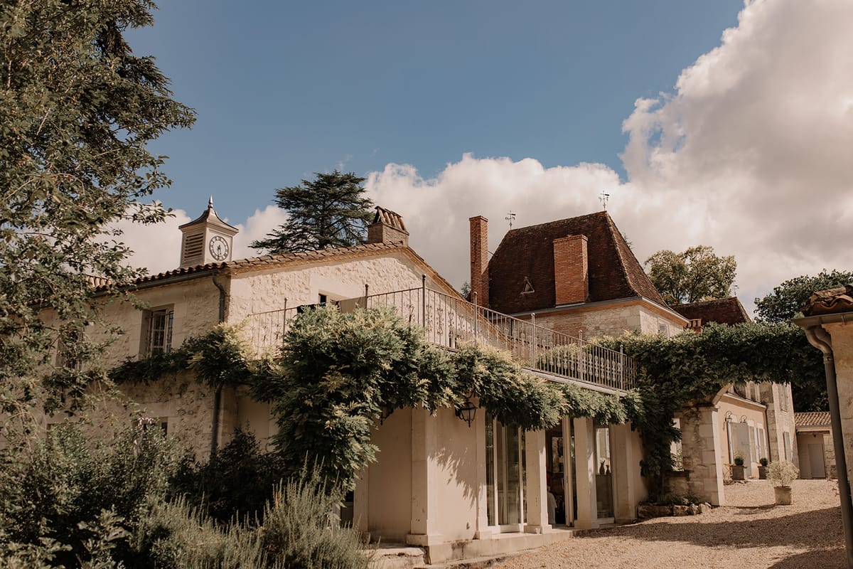 Exterior wide shot of a French country estate featuring cream-toned limestone architecture with terracotta roof tiles, a distinctive clock tower on the left wing, and a wrought-iron first-floor balcony draped with trained climbing greenery. The main building includes a mansard-style turreted roof with brick chimneys and a weather vane, while a ground-floor glazed extension opens onto a gravel courtyard. A secondary outbuilding with pale blue shutters is visible to the right. No people are present in the frame. Potential venue feature image.