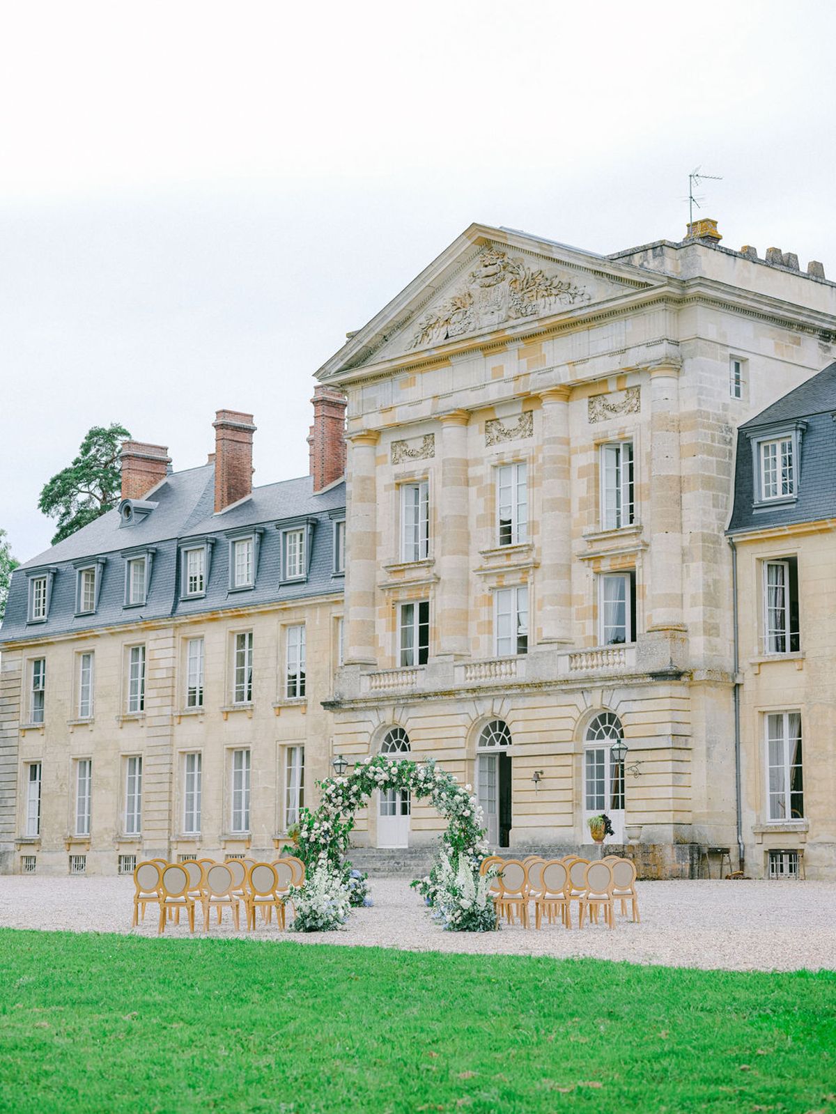 An outdoor ceremony setup is positioned directly in front of a large French classical château, shot from a wide-angle perspective across the lawn. The ceremony area features two sets of gold-framed medallion chairs arranged in rows on either side of a central aisle on a gravel courtyard. A circular floral arch stands at the altar position, constructed with white and blush blooms alongside abundant greenery and white trailing flowers at its base. The château itself is a three-story cream limestone neoclassical building with mansard slate roofs, decorative pediment with carved relief, pilasters, arched ground-floor windows, and brick chimneys — no guests or wedding party are present in the image. The overall styling is classic and refined, with the gold chairs and soft white floral palette complementing the pale stone architecture. Potential venue feature image.
