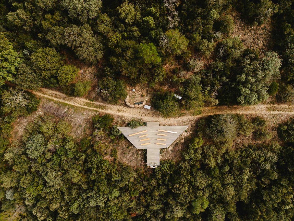 Aerial drone shot of an outdoor wedding ceremony setup in a forest clearing. A triangular wooden deck platform serves as the ceremony space, with two rows of natural wood log benches arranged on either side of a central aisle. A couple — likely the bride and groom — is visible at the narrow end of the platform, facing what appears to be an officiant. A dirt path runs horizontally across the frame, cutting through dense tree coverage, with a parked white vehicle visible on the right side. The ceremony aesthetic is rustic and nature-forward, with raw wood furnishings and no visible floral or fabric decor from this altitude. The composition is a top-down aerial wide shot. Potential venue feature image.