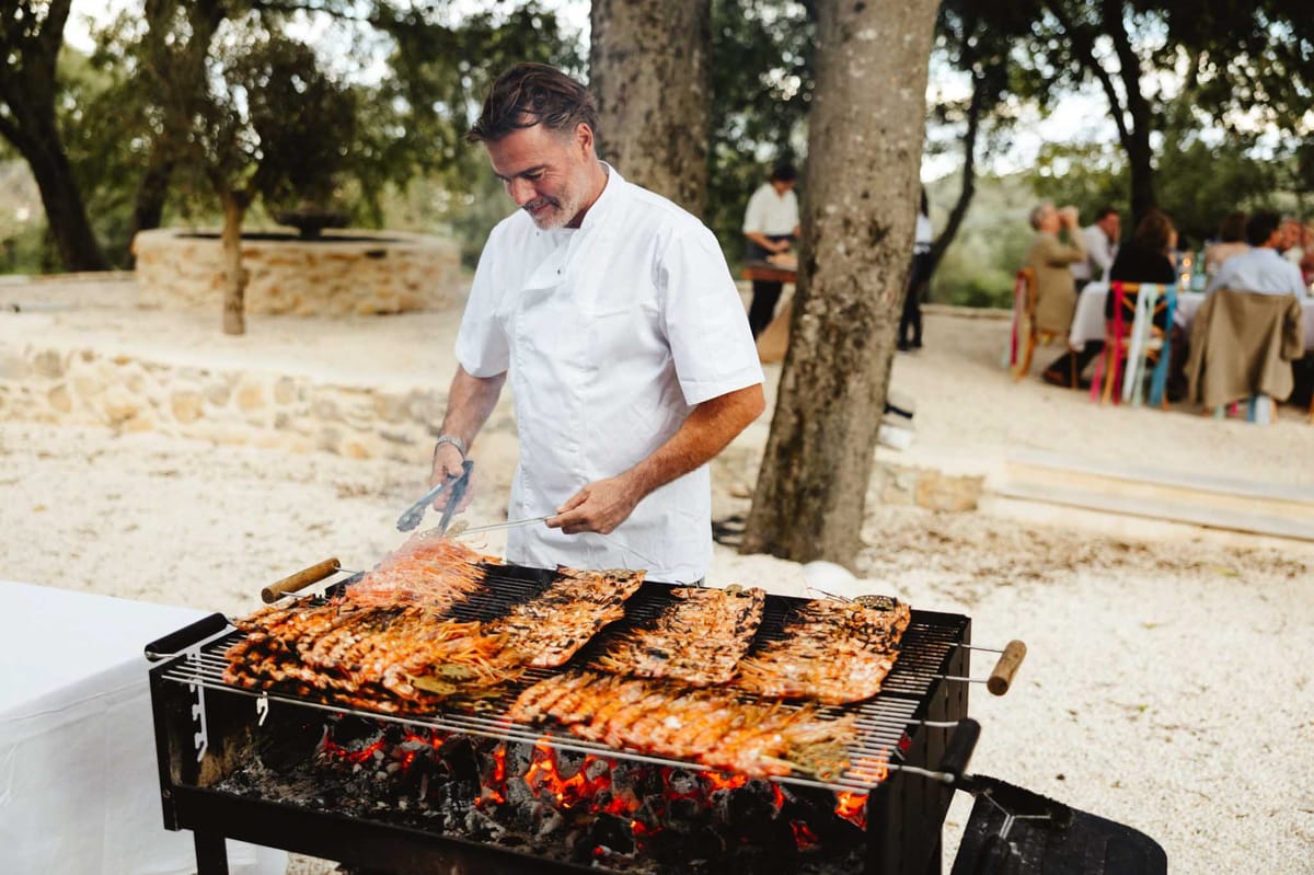 An outdoor wedding reception catering scene at what appears to be a rustic Provençal estate or domaine, with a chef in a white chef's jacket grilling large quantities of whole prawns or langoustines over an open charcoal barbecue with visible glowing embers. The chef is smiling and using metal tongs to tend to the seafood, which covers the entire grill surface. In the background, wedding guests are seated at long tables with natural linen table coverings and colorful chairs — including what appear to be brightly colored bistro-style chairs in yellow, red, and blue — set among mature trees on a gravel and sandy ground. A stone well or water feature is visible to the left. The setting is casual and festive with a relaxed, rustic outdoor dining style. Medium shot with the chef and grill in sharp focus and guests softly blurred in the background.