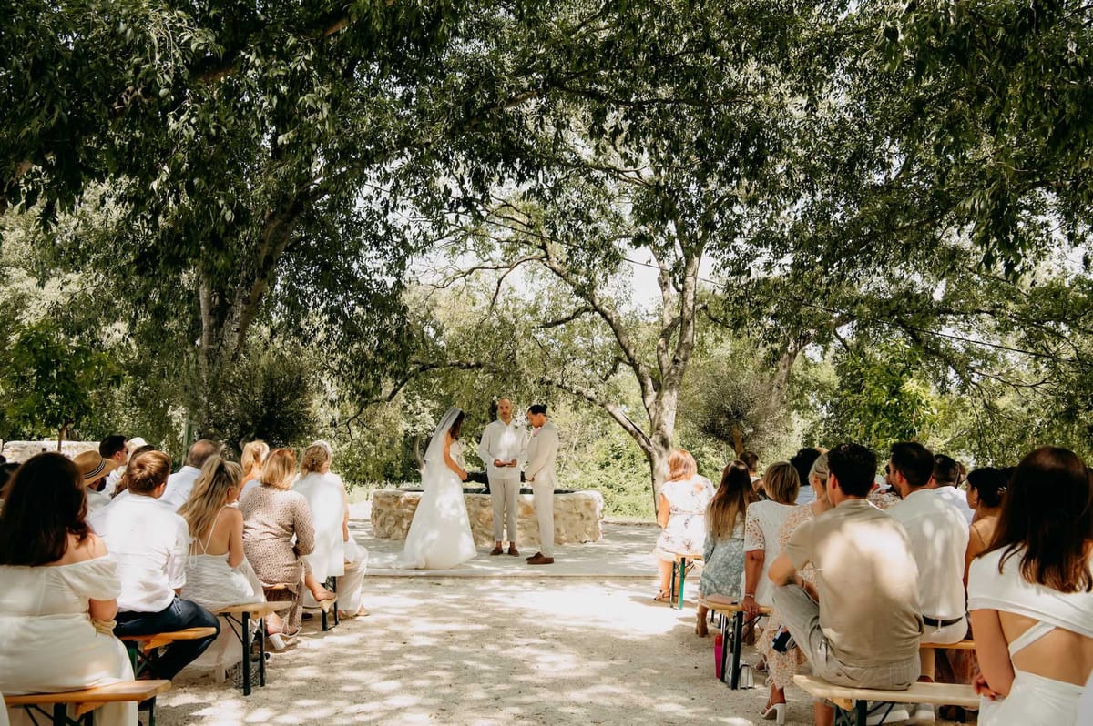 An outdoor wedding ceremony taking place beneath a canopy of mature trees, with dappled sunlight filtering through the foliage onto a gravel or sandy aisle. The bride, wearing a white gown and a long veil, stands facing the groom, who is dressed in a light beige suit, as an officiant stands between them at the far end of the aisle. Approximately 30–40 guests are seated on wooden benches arranged in two rows on either side of the aisle, dressed predominantly in white, cream, and light neutral tones consistent with a warm-weather dress code. The ceremony setting has a relaxed, natural, rustic feel with no visible floral arch or decorative structures, relying entirely on the tree canopy as a natural frame; the overall aesthetic is casual and bohemian. Wide shot taken from the back of the aisle, capturing the full ceremony space and guest seating.