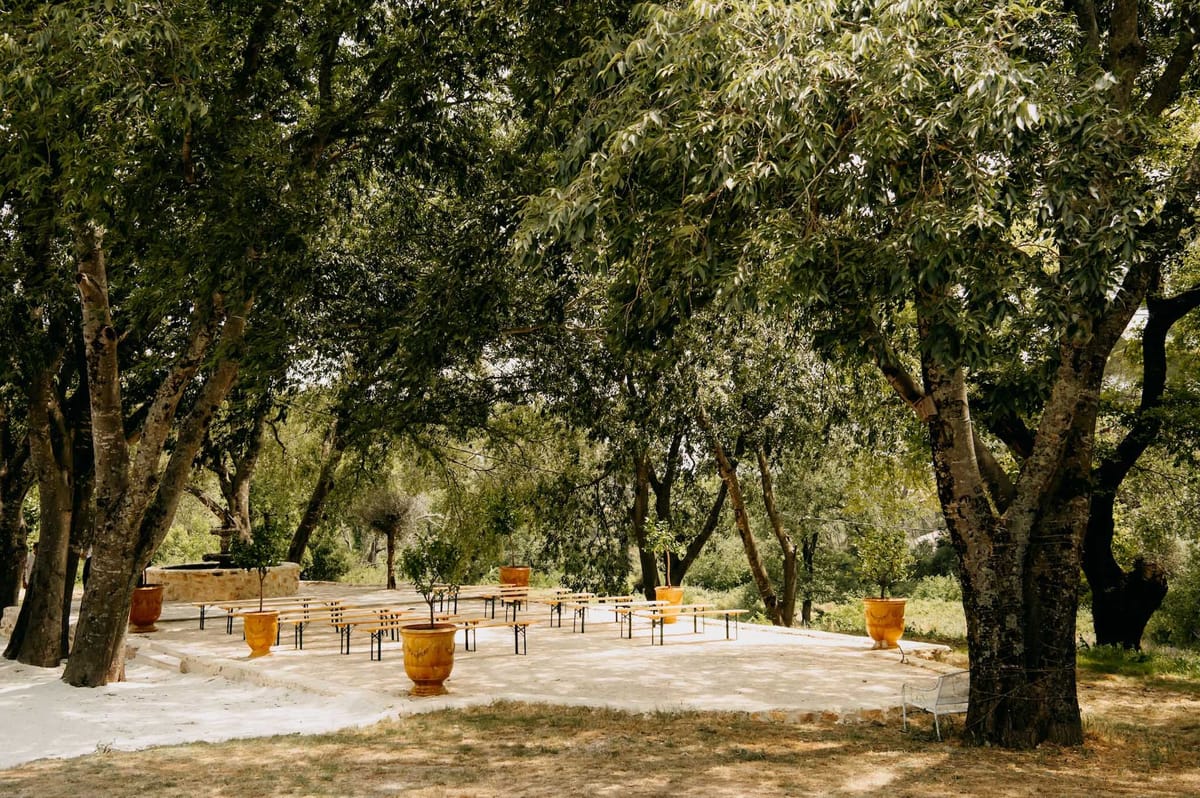 An outdoor ceremony or reception space set up on a paved stone terrace surrounded by mature trees providing natural shade. The space features several rows of dark metal folding benches arranged in a casual, open layout, interspersed with large terracotta-orange Anduze-style ceramic pots containing small potted trees. A low stone structure, likely a fountain or decorative element, is visible at the far left. The overall styling is rustic Provençal, with the warm amber tones of the pots contrasting against the pale stone paving. The image is a wide environmental shot taken from ground level at the edge of the terrace. No people are present. Potential venue feature image.