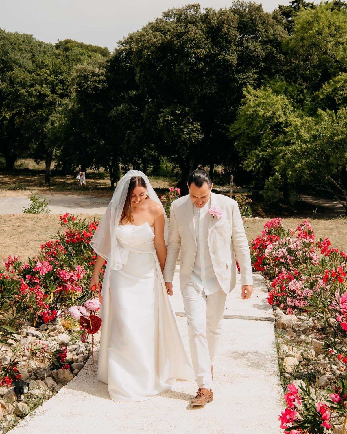 The bride and groom walk hand-in-hand along a sunlit stone pathway in an outdoor garden setting, both smiling downward in a candid, relaxed moment. The bride wears a strapless ivory satin column gown with a fingertip-length veil, while the groom is dressed in a cream linen suit with white trousers, tan loafers, and a pink peony boutonniere. The pathway is flanked by low stone-edged garden beds planted with deep red and hot pink oleander blooms, along with what appears to be a red anthurium visible at lower left. A second figure, likely a guest or child, is faintly visible in the background among mature trees. This is a full-length portrait shot taken in bright midday natural light with a modern, relaxed outdoor style.
