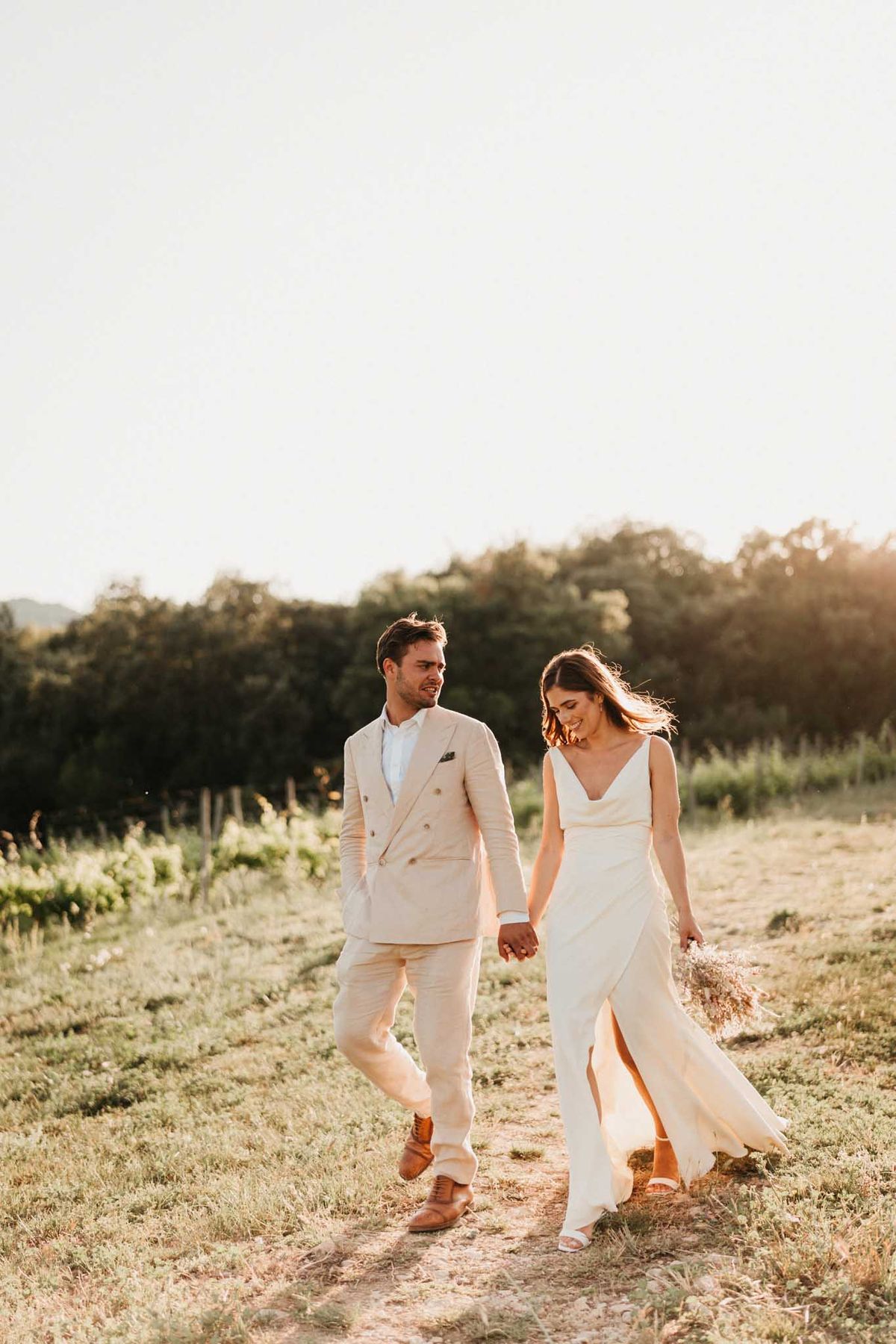 Couple walking vineyard path at golden hour, groom in cream linen suit, bride in ivory cowl-neck slip dress