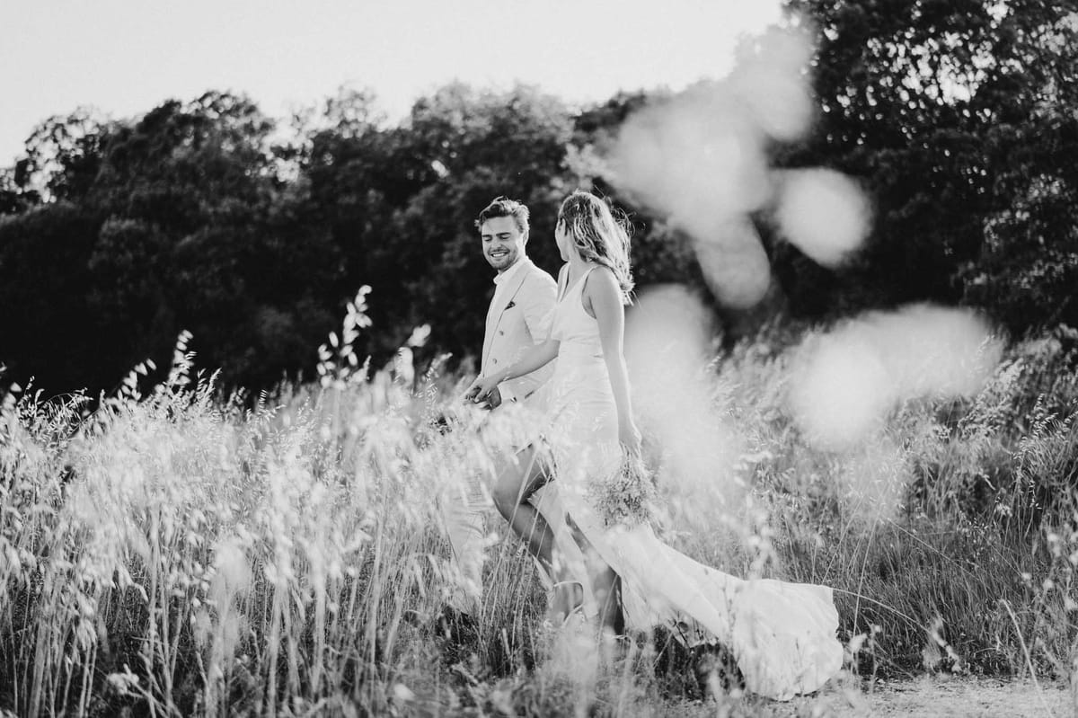 Black-and-white portrait of bride and groom walking hand-in-hand through tall grass at golden hour