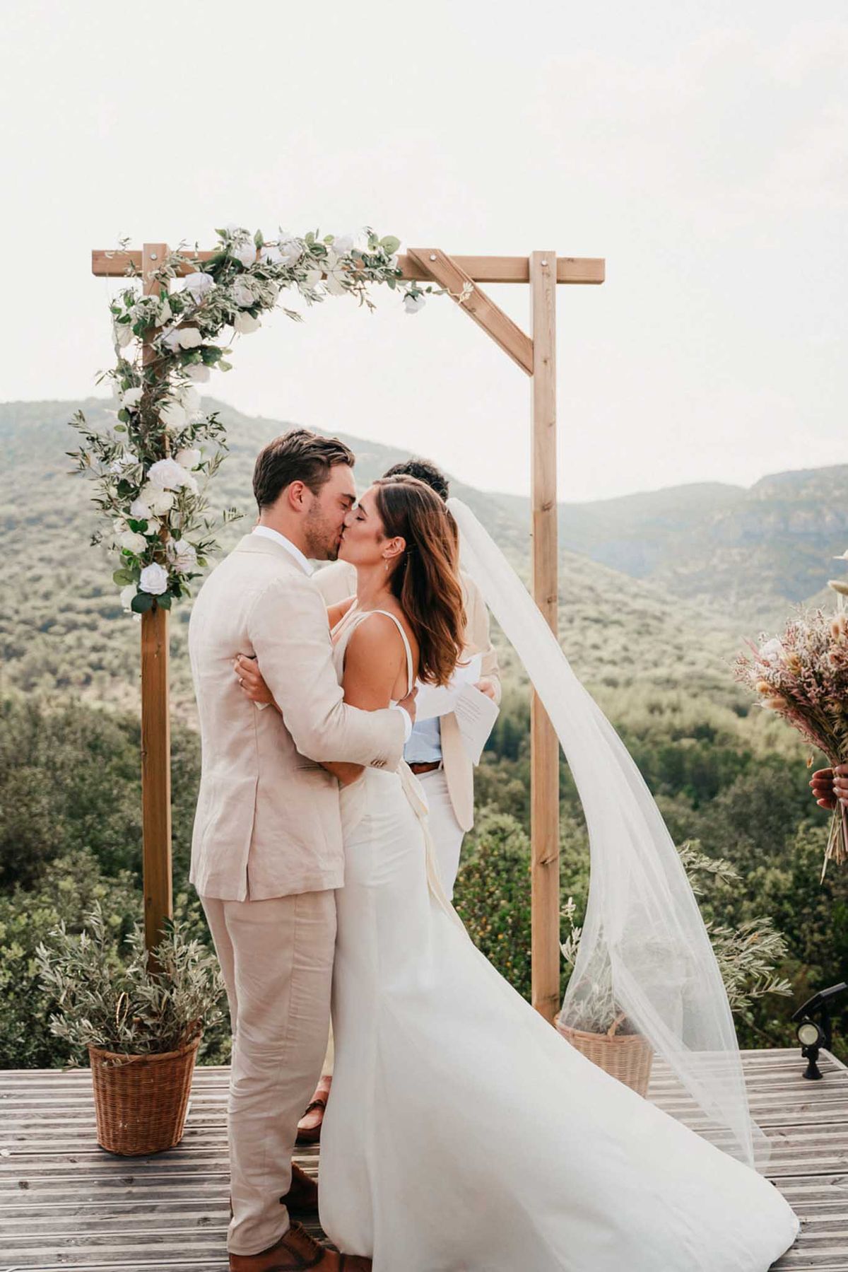 First kiss under wooden arch with white roses and eucalyptus, bride in gloves and flowing cathedral veil