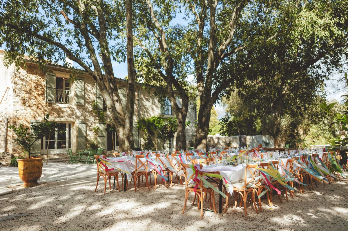 An outdoor wedding reception setup in the courtyard of a French stone mas or bastide, featuring long banquet-style tables arranged in a U-shape on a gravel surface beneath large mature trees. The tables are dressed with white linens and set with glassware, and the natural wood cross-back chairs are decorated with multicolored ribbon streamers in pink, yellow, green, and blue, creating a festive, playful decor palette. The setting is daytime with dappled natural light filtering through the tree canopy, and the stone building in the background has sage green shutters and French doors, with terracotta planters flanking the entrance. Wide shot capturing the full table arrangement and venue courtyard. Potential venue feature image.