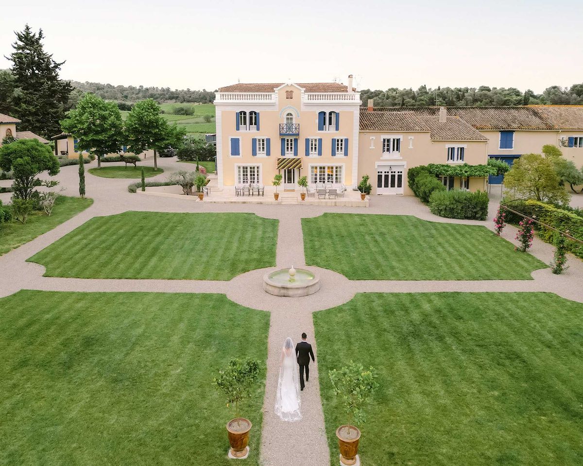 An aerial drone shot captures a bride and groom walking hand in hand along a gravel pathway leading toward a French country manor. The bride wears a white gown with a long cathedral-length veil, and the groom is dressed in a black suit. The formal garden features symmetrically manicured lawn panels divided by gravel paths, with a central stone fountain and terracotta potted topiaries flanking the walkway. The manor itself is a three-storey peach-rendered building with blue shutters, white trim, and a terracotta tile roof, flanked by lower annexe buildings in a matching style; outdoor seating with bistro-style furniture is arranged on the front terrace. Potential venue feature image.