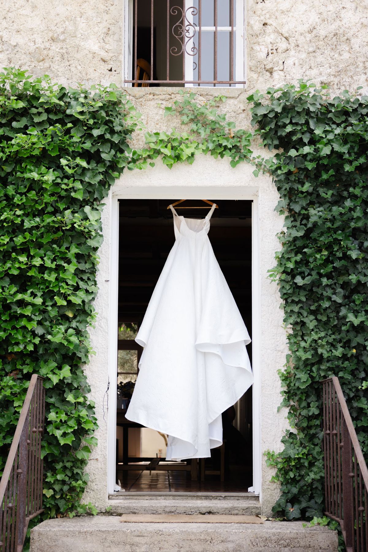 A getting-ready detail shot showing a white wedding dress hanging from a wooden hanger in an open doorway of a French stone building. The dress features thin straps, a fitted bodice, and a layered, full skirt with tiered ruffles in a clean white fabric. The doorway is framed by ivy climbing the exterior walls on both sides, and a wrought-iron railing borders the entrance steps. An upper window with decorative iron bars is visible above. The composition is a centered wide shot, shot from the exterior looking toward the darkened interior of the building.