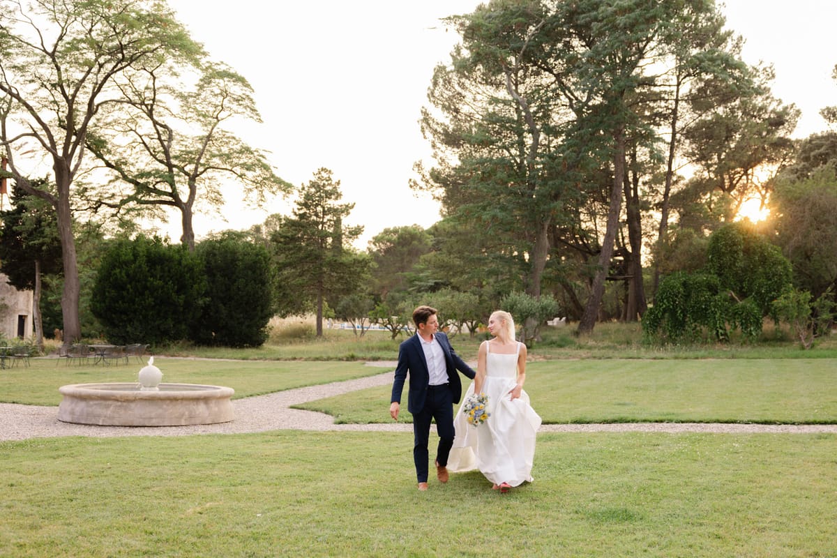 Bride and groom walking across lawn at golden hour at French domaine with fountain and garden furniture