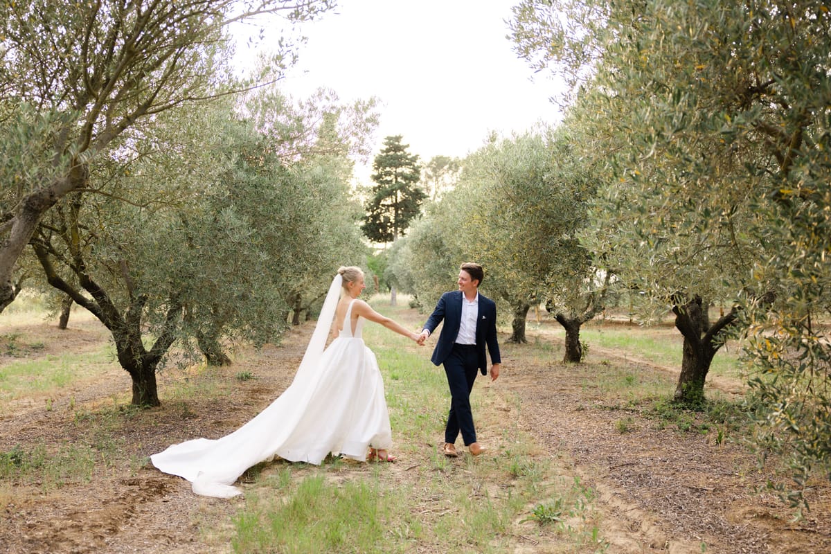 Bride in white ball gown with cathedral veil leading groom in navy suit through an olive grove path