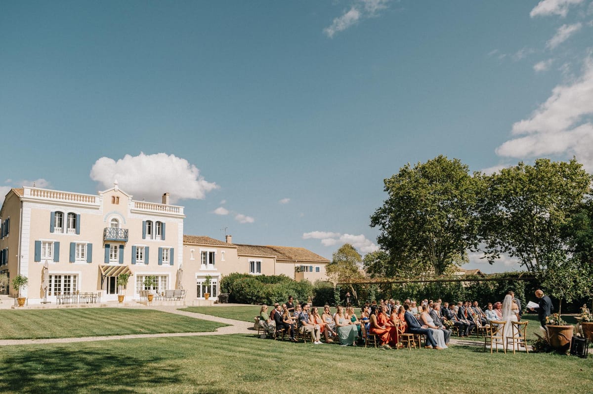 Outdoor wedding ceremony on the lawn of a peach-toned French manor with blue shutters and seated guests