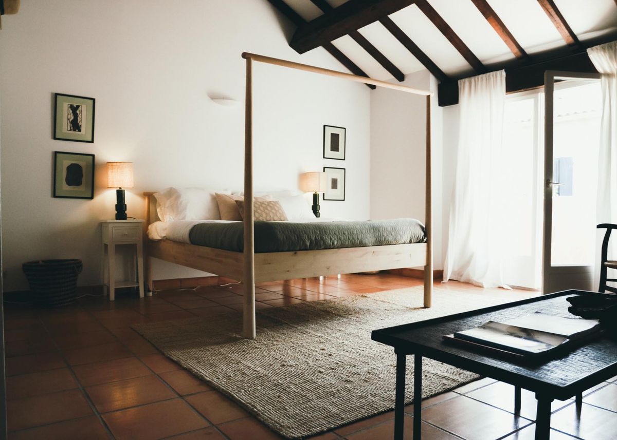 This image shows a hotel or venue bedroom interior rather than a wedding photograph. The room features a minimalist natural wood four-poster bed frame dressed with olive green and white linen bedding, positioned on terracotta tile floors over a woven natural fiber rug. The vaulted ceiling has exposed dark wood beams, white walls display framed abstract artwork, and lighting includes two black-based table lamps with warm-toned shades. A dark wood low table is visible in the foreground, white sheer curtains frame a open glass door letting in natural light, and a small white bedside table sits to the left. The overall decor palette is warm and minimal, combining natural wood, terracotta, and muted neutrals. Wide interior shot. Potential venue feature image.