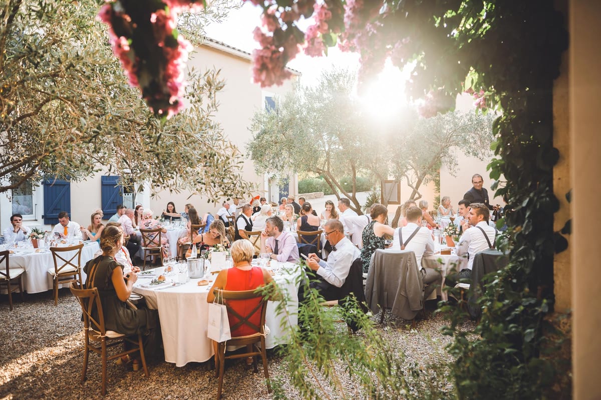 Wedding reception dinner in Provencal courtyard with round tables, cross-back chairs and golden backlight through archway