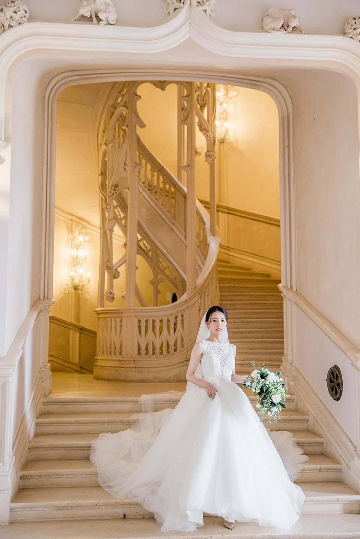 Bride in white ball gown with cathedral veil and white rose bouquet on ornate marble staircase inside French chateau