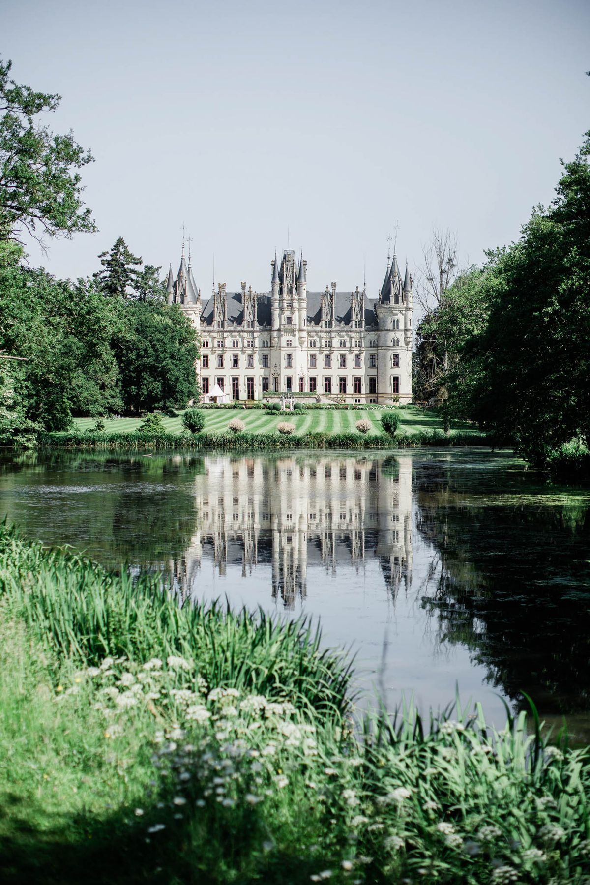 A wide-shot exterior view of a large French château with Neo-Gothic architecture, featuring pointed turrets, ornate stone façade, and steep slate rooflines. The building is photographed from across a still ornamental lake, with its full reflection visible in the calm water. A white marquee or pavilion tent is partially visible on the manicured striped lawn in front of the château, suggesting wedding event setup. The foreground includes wild grasses and small white flowers along the water's edge. No people are clearly identifiable at this distance. Potential venue feature image.