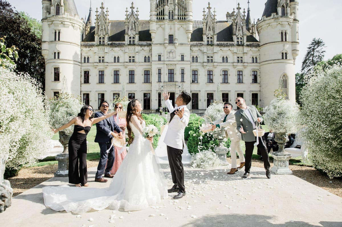 A post-ceremony confetti or petal toss moment taking place on the gravel forecourt of a large French Gothic-style château, with white limestone towers and ornate stone detailing visible across the full width of the frame. The bride wears a white ball gown with a full tulle skirt and long train, holding a white bouquet, while the groom wears a white tuxedo jacket with black trousers. Six guests surround them — dressed in black, navy, blush, cream, and sage outfits — laughing and tossing white flower petals in the air. The ceremony aisle is flanked by large stone urns overflowing with white gypsophila arrangements, with white petals scattered across the ground. The overall decor palette is white and ivory with a classic, formal style. Wide shot with the château facade centered in the background. Potential venue feature image.