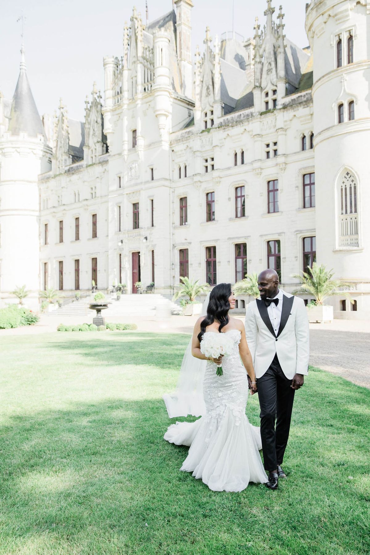 Couple walking hand in hand on lawn before Gothic-Revival chateau with spires and fountain