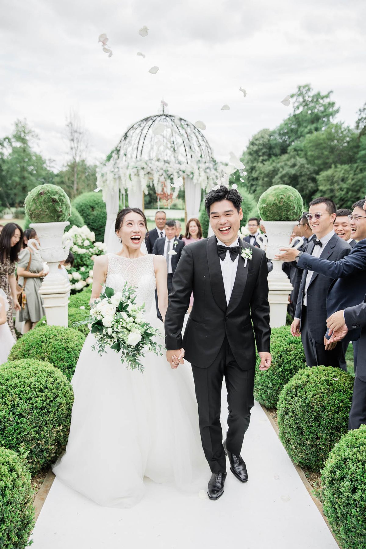 Bride and groom walk down the aisle laughing as guests toss rose petals in a formal French garden