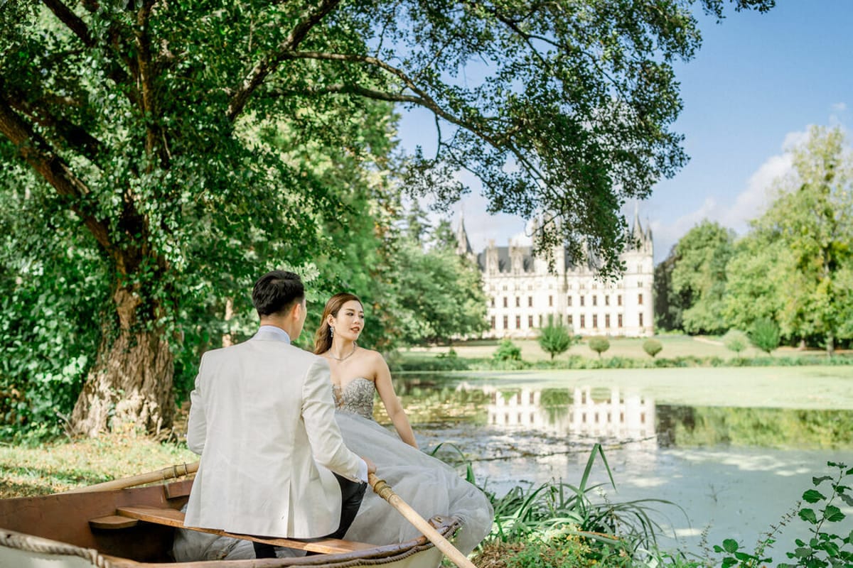 A couple portrait shot outdoors, with the bride and groom seated together in a small wooden rowboat on a calm pond. The groom, shown from behind, wears a white dinner jacket with dark trousers, while the bride faces him wearing a strapless silver-grey ball gown with embroidered bodice detailing, paired with drop earrings and a delicate necklace. In the background, a large white French château with pointed turrets and ornate architecture is reflected in the still water of the pond. The composition is a medium-wide shot taken from the shoreline, framing the couple under overhanging tree branches. Potential venue feature image.