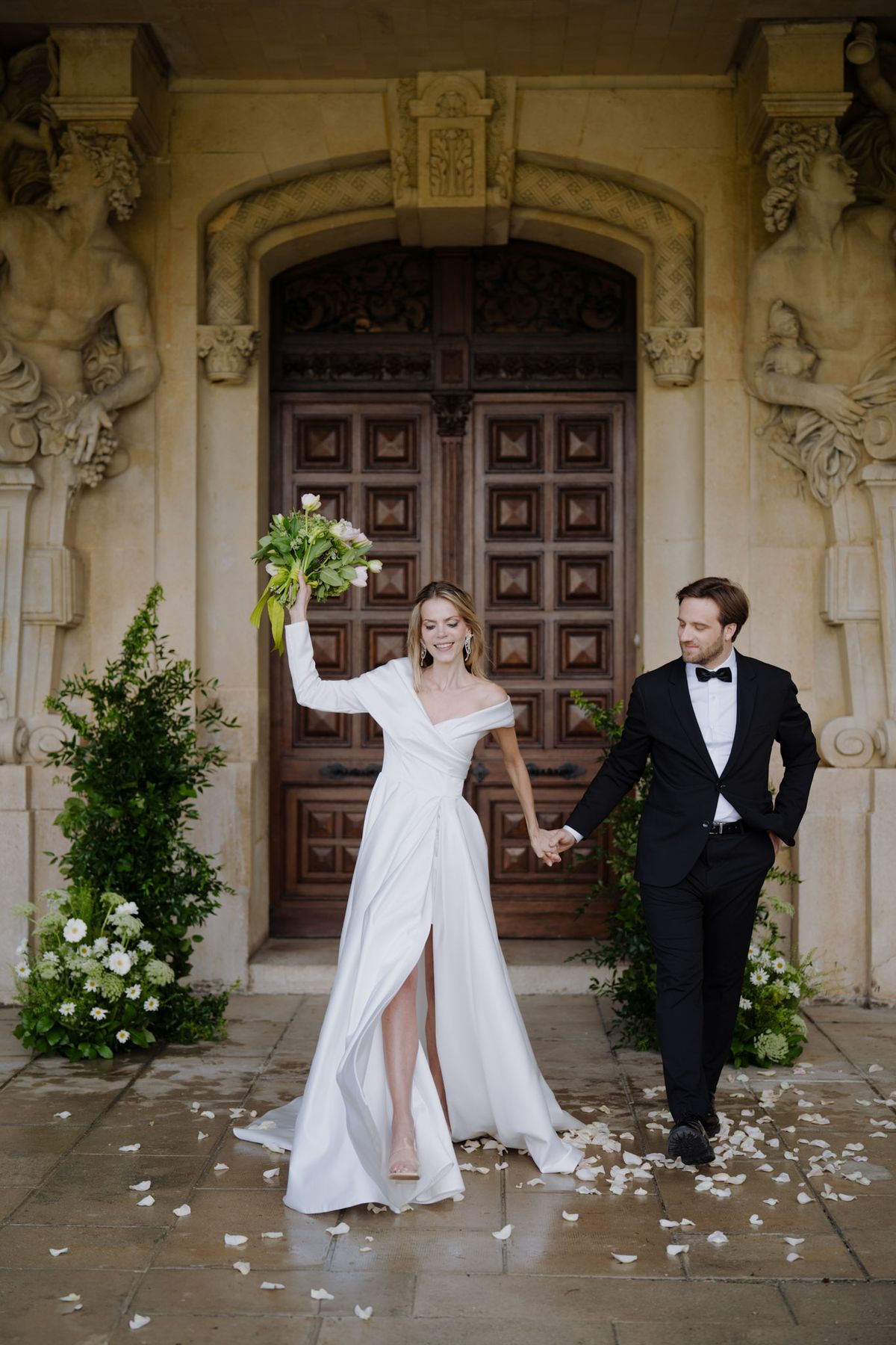 Bride in one-shoulder gown and groom in tuxedo walking past ornate stone doorway with scattered petals