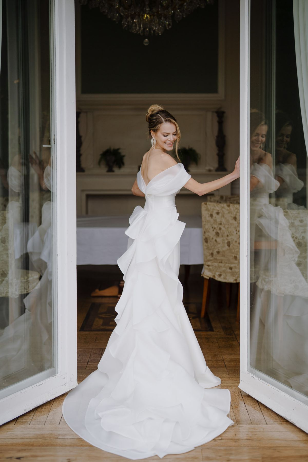 Bride looking over shoulder in off-shoulder ruffle gown at chateau French doors with crystal chandelier