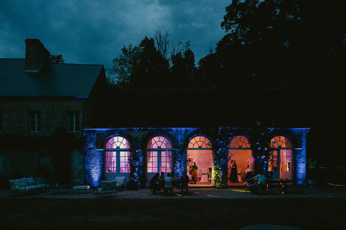 Night shot of chateau orangerie with blue uplighting, pink interior glow, and guests outside