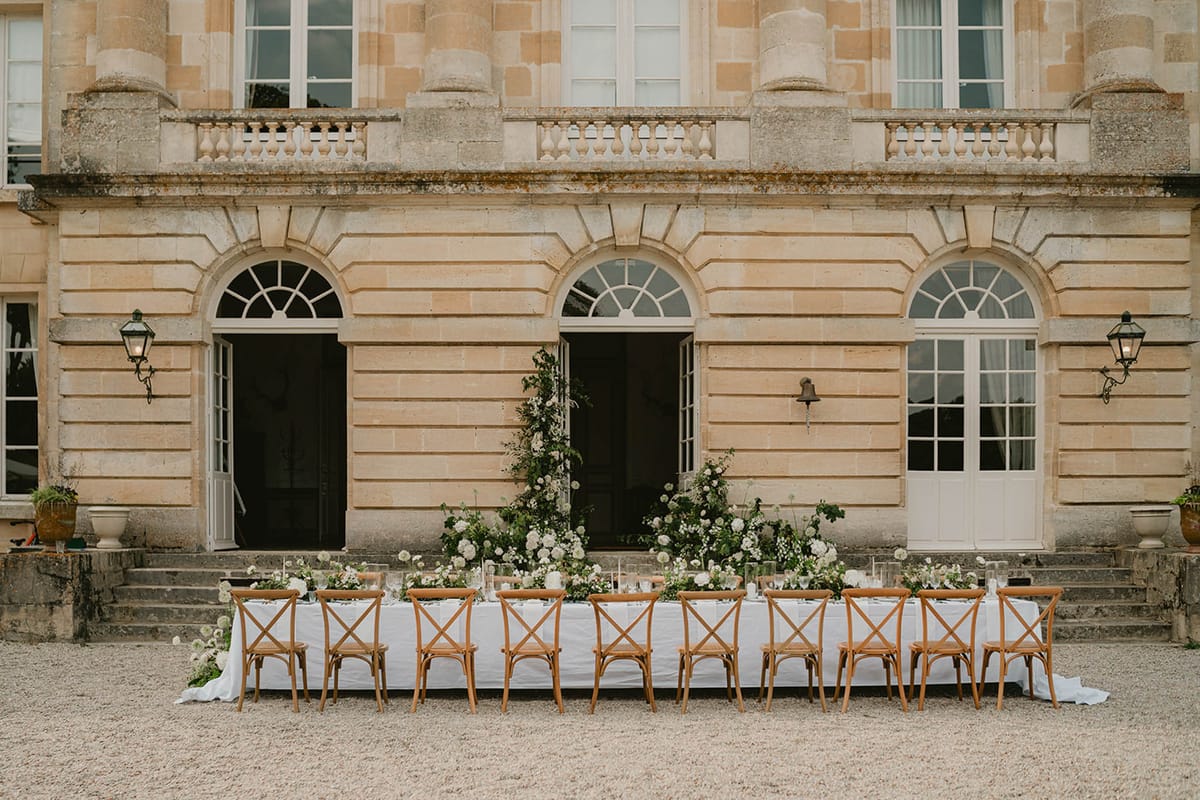 Long table with white rose and allium runner and tall floral installation before chateau with lanterns
