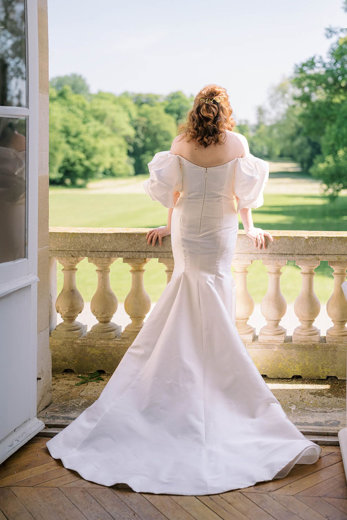 Bride from behind on chateau terrace in off-shoulder mermaid gown with cathedral train overlooking gardens
