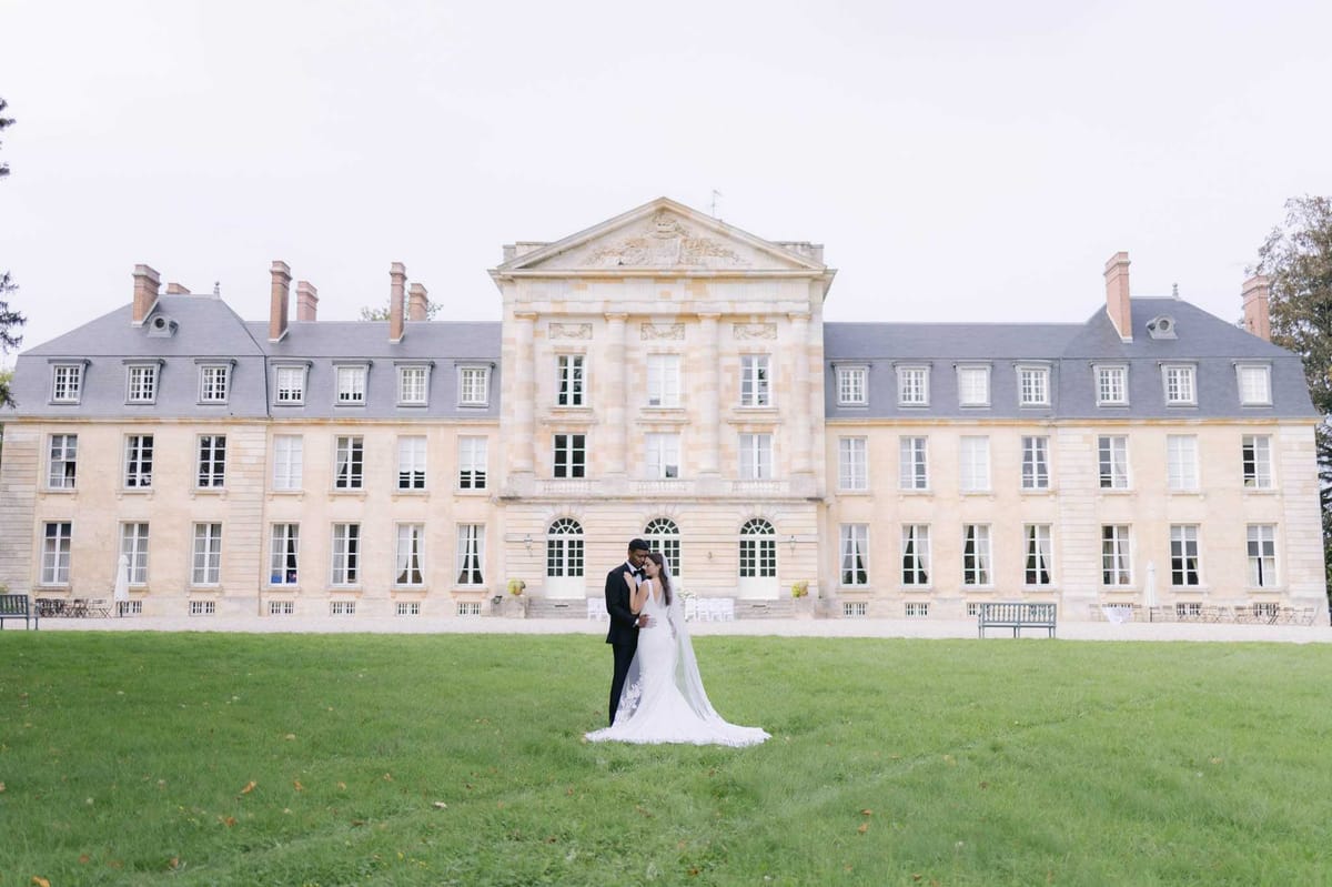 Couple touching foreheads with cathedral veil on lawn before symmetrical classical limestone chateau