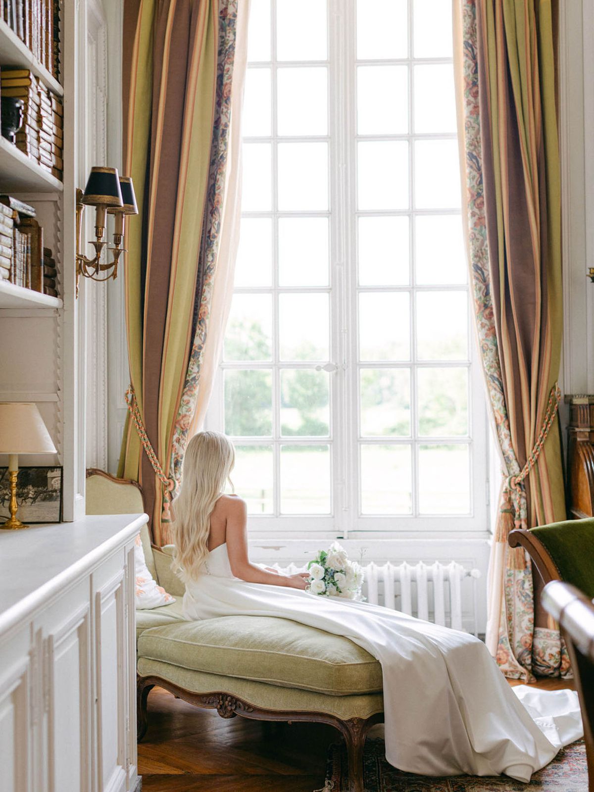 Bride from behind on sage chaise longue with white rose bouquet facing window in chateau library