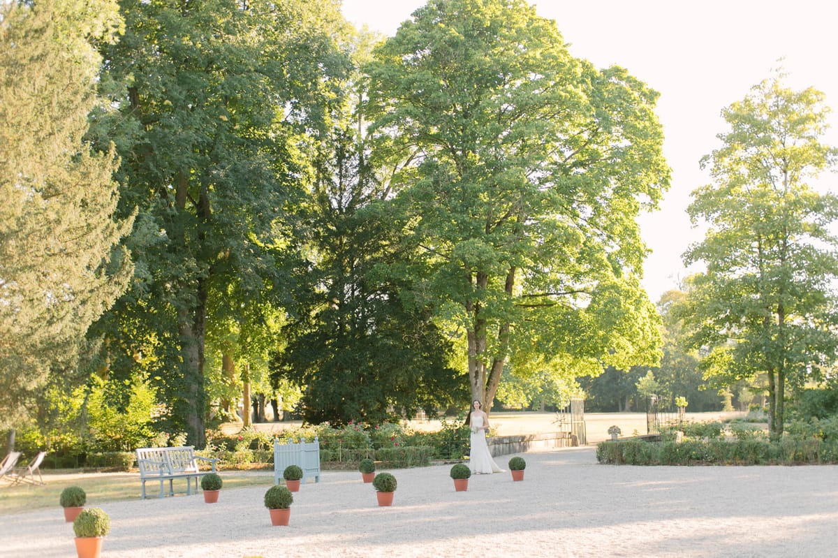 A bride stands alone in the grounds of a French estate during golden hour, positioned in the middle distance against a backdrop of large mature trees. She is wearing a fitted white gown and holding a bridal bouquet, appearing small within the wide landscape composition. The gravel courtyard is lined with clipped boxwood topiaries in terracotta pots, and powder blue painted wooden benches are placed along the path. Flowering rose beds and neatly trimmed hedging border the space, and a decorative iron structure is visible further along the gravel path to the right. The wide shot captures the full scale of the formal garden grounds in warm afternoon light.