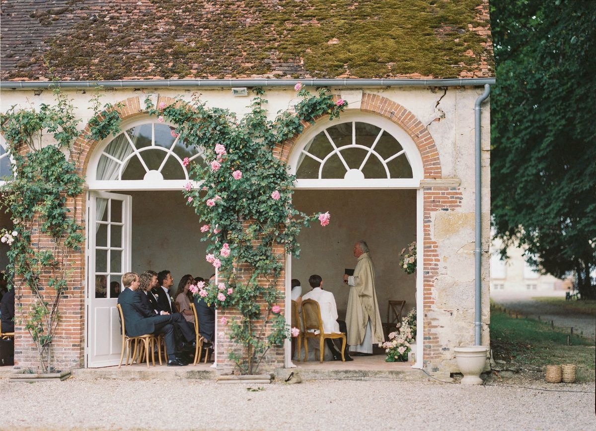 Wedding ceremony inside open-arched chateau outbuilding with climbing roses and seated guests on gold chairs