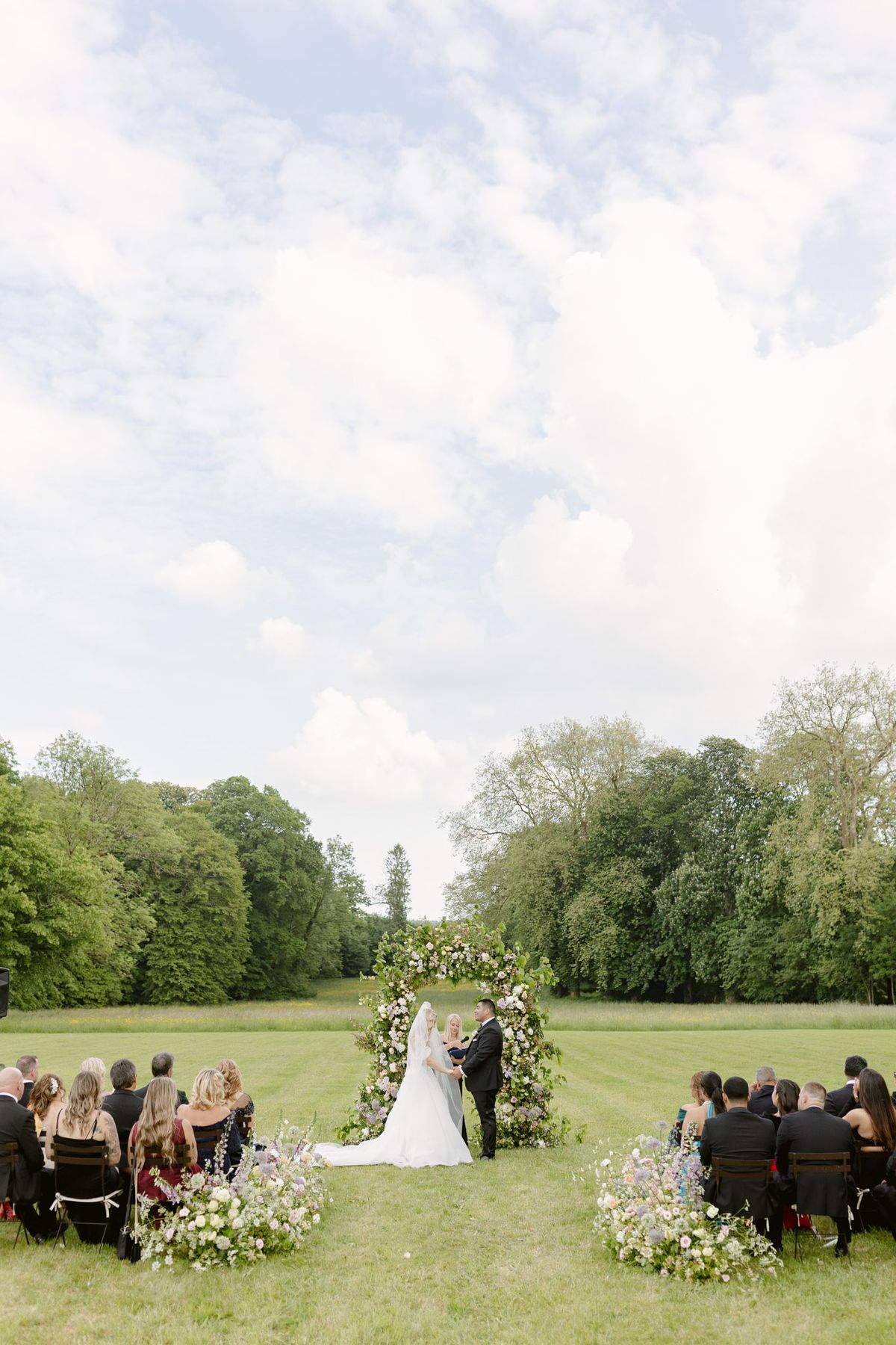 Outdoor ceremony with couple under circular floral arch in meadow, guests seated in wooden chairs on either side