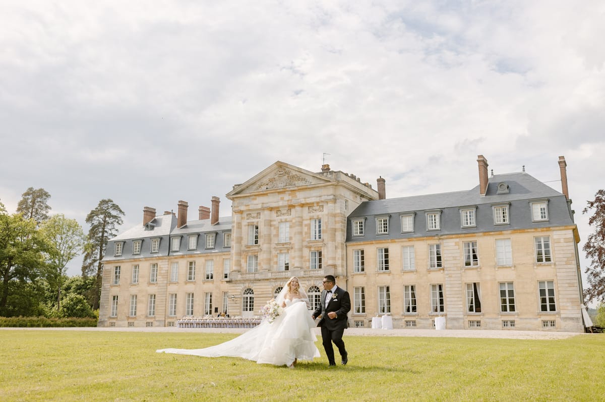 Bride in white ball gown with cathedral train and groom in black tuxedo walking across lawn before neoclassical chateau