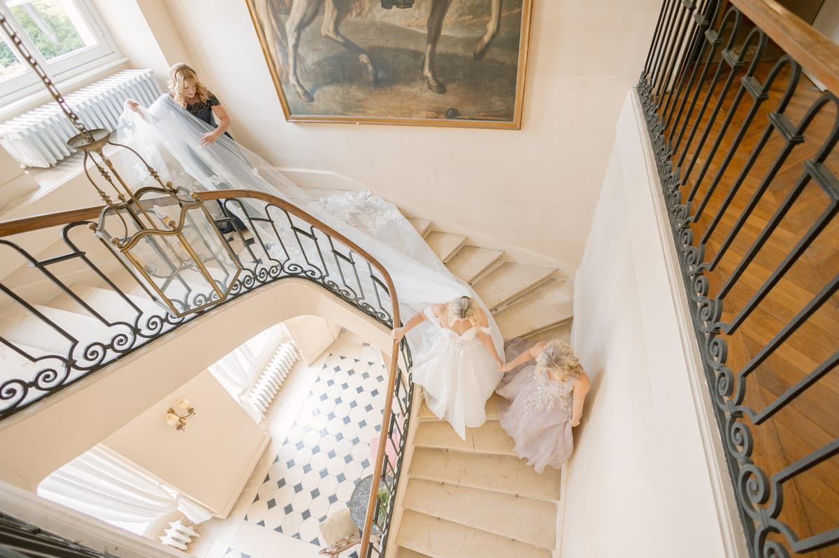 An overhead wide shot capturing the bride descending a grand curved interior staircase inside a château, accompanied by two women — one walking closely beside her and one above on the stairs lifting and managing the long cathedral-length white veil with lace trim. The bride wears a white ballgown with a lace-detailed back and her hair is pinned up. The woman beside her wears a blush-toned embellished gown, while the woman handling the veil above wears a dark navy or black short-sleeved dress. The staircase features ornate wrought-iron and brass scrollwork railings with stone steps, and the entry hall below has a classic black-and-white diamond-patterned tile floor visible from above. A large antique oil painting depicting a horse hangs on the landing wall. The composition is shot from directly above, emphasizing the sweep of the veil cascading down the stairs. Potential venue feature image.