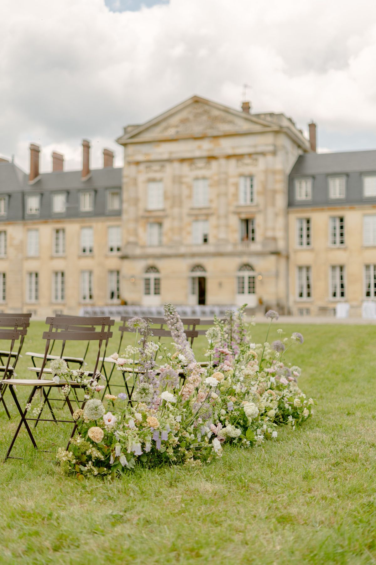 An outdoor wedding ceremony setup on the grounds of a large French château, photographed from ground level with the focus on a loose floral ground arrangement placed at the end of a ceremony aisle. The floral installation is composed of peach garden roses, lavender and lilac delphiniums, white ranunculus, mauve sweet peas, allium globes, and trailing greenery arranged in a natural, unstructured style directly on the lawn. Dark brown folding metal chairs are lined up in rows along the aisle, with additional rows of white chairs visible further back near the château. The background features a grand stone classical-style French château with a symmetrical façade, pediment detail, and mansard roof, rendered softly out of focus. The overall decor palette is soft pastel — blush, lavender, ivory, and peach — with a romantic garden-style aesthetic. Wide shot with shallow depth of field emphasizing the foreground floral detail. Potential venue feature image.