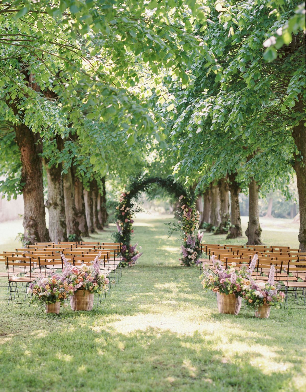 Outdoor ceremony aisle with wooden chairs beneath tree-lined allee leading to circular floral arch in peach and mauve
