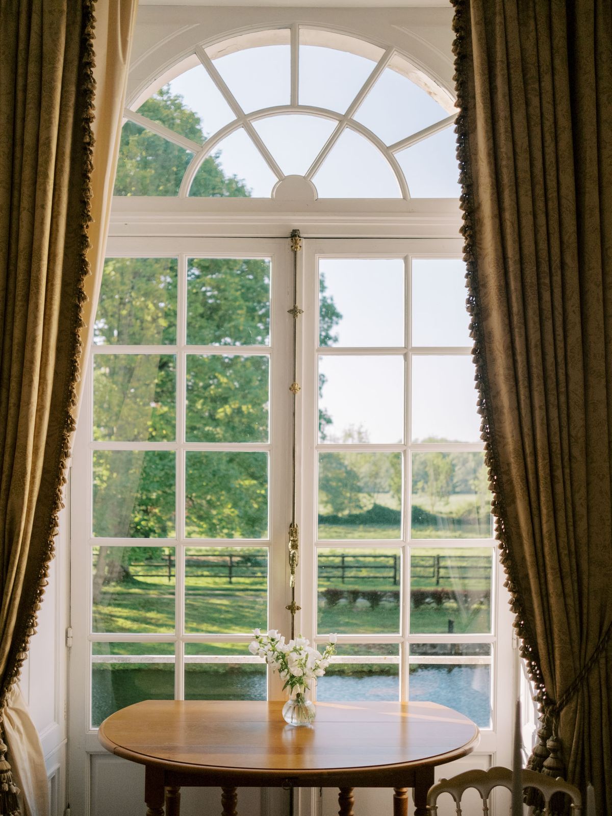 Arched French window with olive green velvet curtains and white sweet peas on side table overlooking manicured grounds