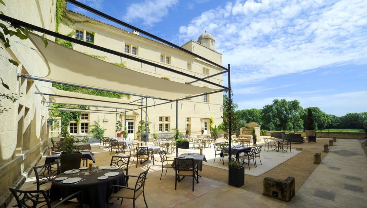 Wide-angle exterior shot of a large French chateau terrace set up for an outdoor reception or dining event. The terrace features multiple round and rectangular tables dressed in dark charcoal and black tablecloths with white plate settings, surrounded by wrought-iron chairs. A large cream-colored sail shade canopy on a black metal frame covers a portion of the terrace. The pale limestone chateau facade, partially covered in climbing ivy, rises several stories and includes a domed turret. Potted topiaries and ornamental plants are arranged around the terrace perimeter, with open parkland and mature trees visible beyond the terrace balustrade. No people are present in the image. Potential venue feature image.