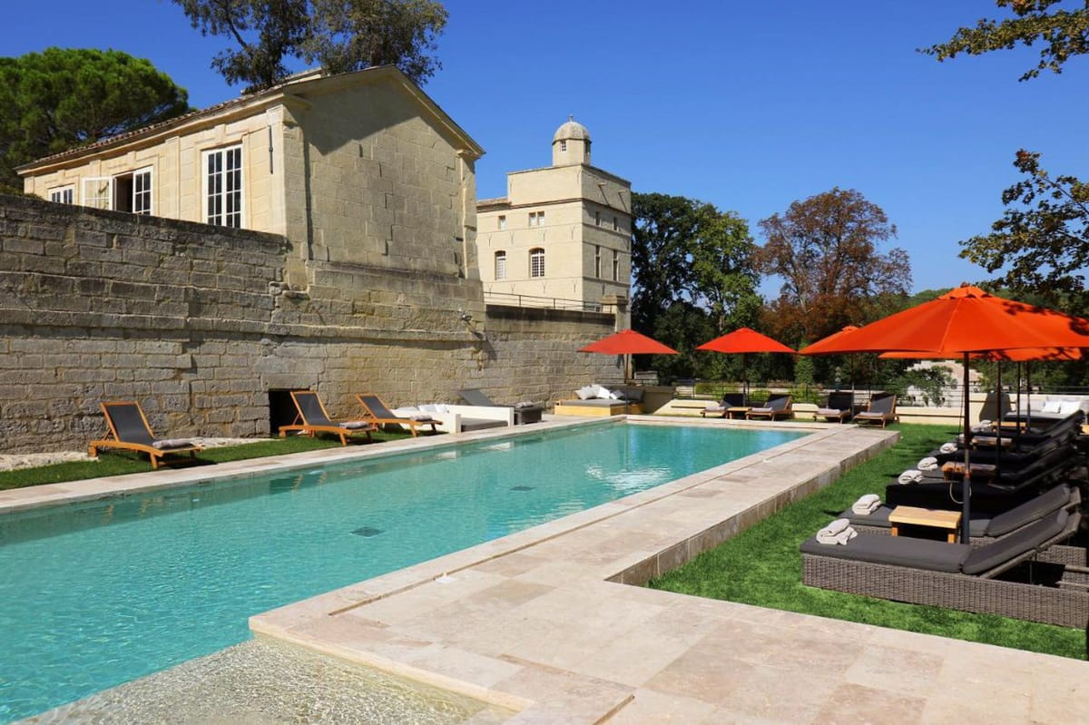 Outdoor pool area at a French château venue, photographed in a wide shot with no people present. The rectangular pool features pale stone coping and terracotta-tinted water, bordered by a limestone terrace. Sun loungers with dark charcoal cushions and teak frames are arranged on both sides of the pool, accompanied by several large burnt-orange parasols. White daybeds are visible against the historic stone retaining wall on the left. The venue buildings are visible in the background — a classical French stone structure with tall white-framed windows and a second taller pavilion with a domed cupola, both constructed from warm beige cut stone. Potential venue feature image.