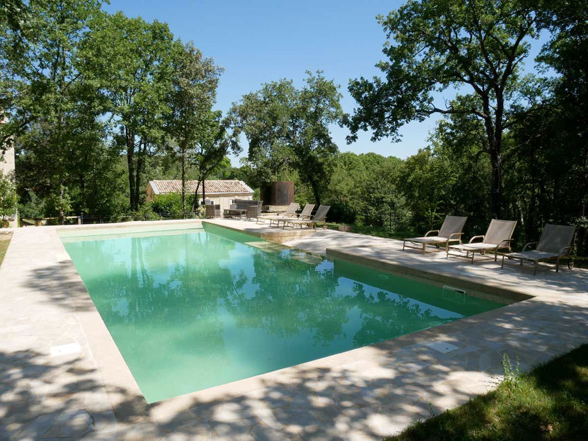 Outdoor venue feature shot showcasing a large rectangular swimming pool with pale turquoise water, surrounded by a wide pale stone terrace. Several tan-colored sun loungers are arranged along the right side and far end of the pool deck. A seating area with lounge chairs is visible in the background near what appears to be a Corten steel cylindrical water feature or sculpture. Stone-roofed outbuildings are partially visible through the trees in the background. No people are present in the image. The overall aesthetic is clean and understated, consistent with a Provençal-style property. Potential venue feature image.