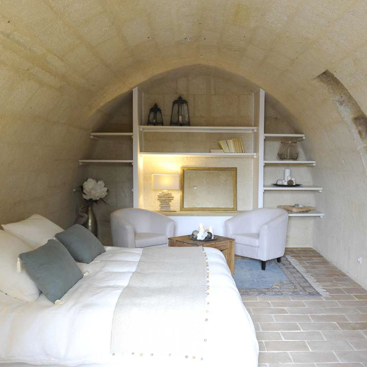 Interior shot of a bedroom suite within a venue featuring a vaulted stone ceiling with an arched alcove, typical of historic French property architecture. The room is styled in a neutral palette of white, cream, and muted sage grey, with a large bed dressed in white linen and grey cushions in the foreground. Two white upholstered tub chairs flank a small wooden coffee table topped with white candles, set on a faded blue and beige area rug over terracotta tile flooring. White open shelving built into the arched alcove displays black lanterns, cream-colored books, a wicker lantern, and decorative objects, with a gold-framed mirror and a lit table lamp adding warm accent lighting. Potential venue feature image.