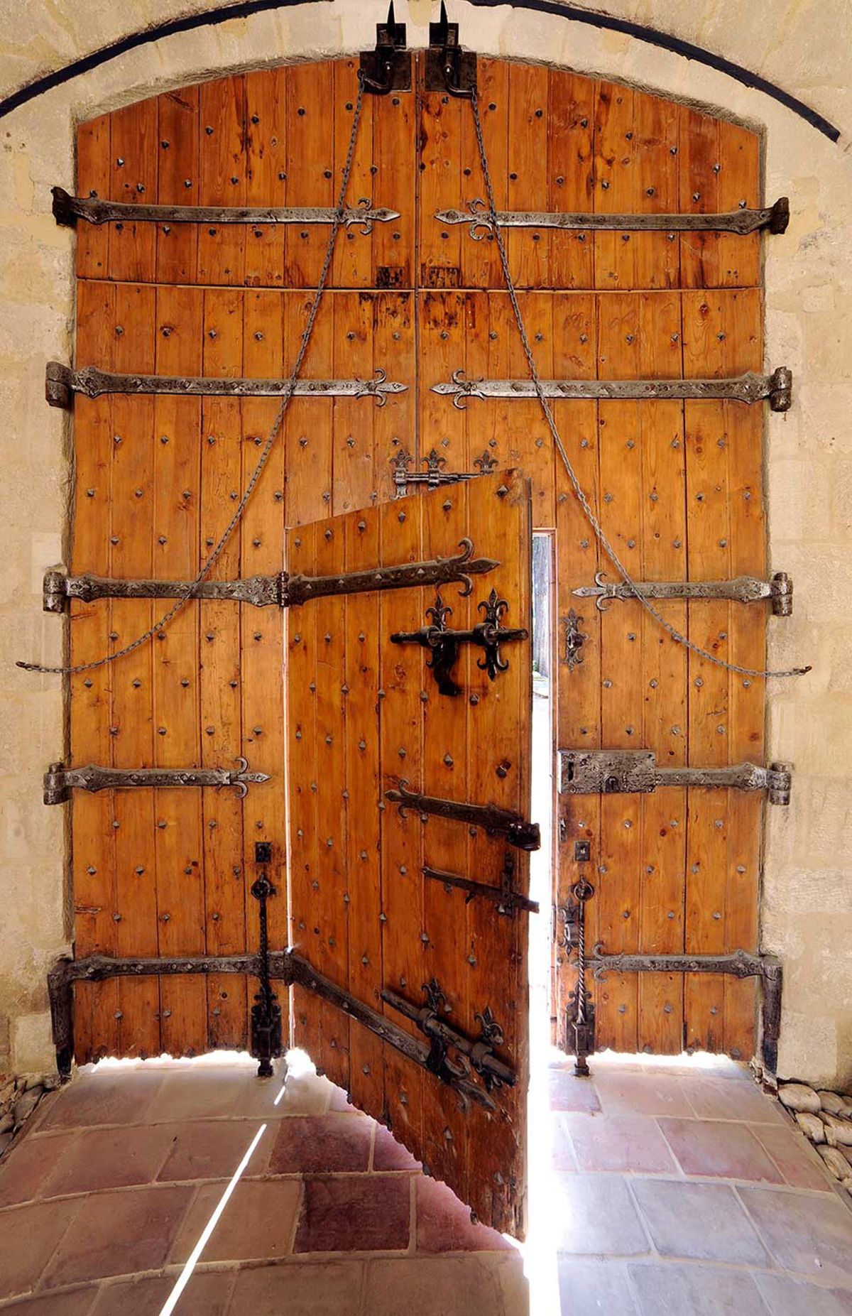 A close-up architectural detail shot of a large set of antique wooden double doors set within a stone archway. The doors are constructed from warm amber-toned vertical planks studded with iron nailheads, and are reinforced with multiple horizontal wrought iron strap hinges featuring decorative fleur-de-lis finials. Iron chains connect the hinges across the door panels. One door is slightly ajar, revealing a glimpse of bright light beyond. The threshold features terracotta and stone tile flooring, and the surrounding archway is built from pale cut limestone blocks. No people are present. Potential venue feature image.
