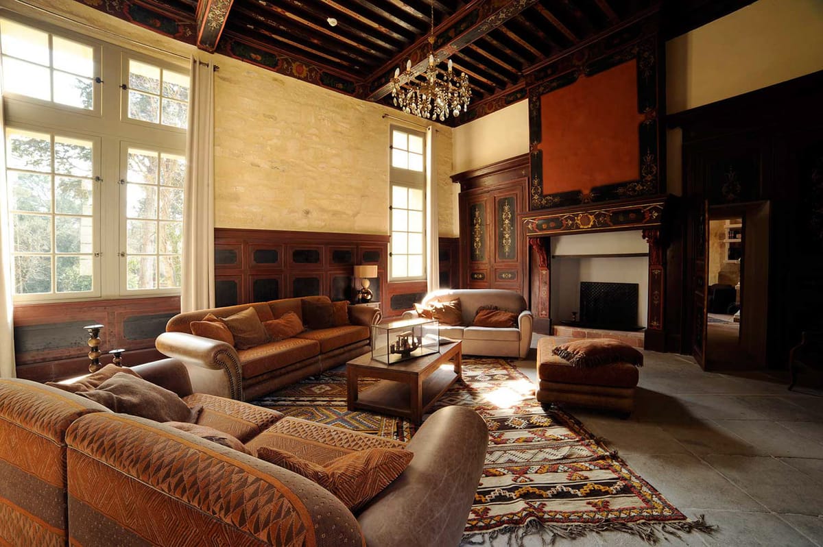 Interior wide shot of a grand salon within a château or historic manor, shown without any wedding guests or couple present. The room features double-height ceilings with dark exposed wooden beams painted with ornate floral and foliate motifs in red, gold, and black. A crystal chandelier hangs centrally from the ceiling. The walls combine exposed limestone with dark wood panelling, also decorated with painted floral patterns, and a large ornate fireplace with an elaborately carved and painted surround anchors the right side of the room. The seating area consists of brown leather and caramel velvet sofas and armchairs arranged around a glass-topped wooden coffee table, all placed on layered Berber-style kilim rugs in cream, red, mustard, and blue geometric patterns. Tall white-framed windows with sheer white curtains line the left wall, allowing warm natural light to stream across the stone-tiled floor. The overall interior style mixes classical French château architecture with warm, eclectic furnishings. Potential venue feature image.