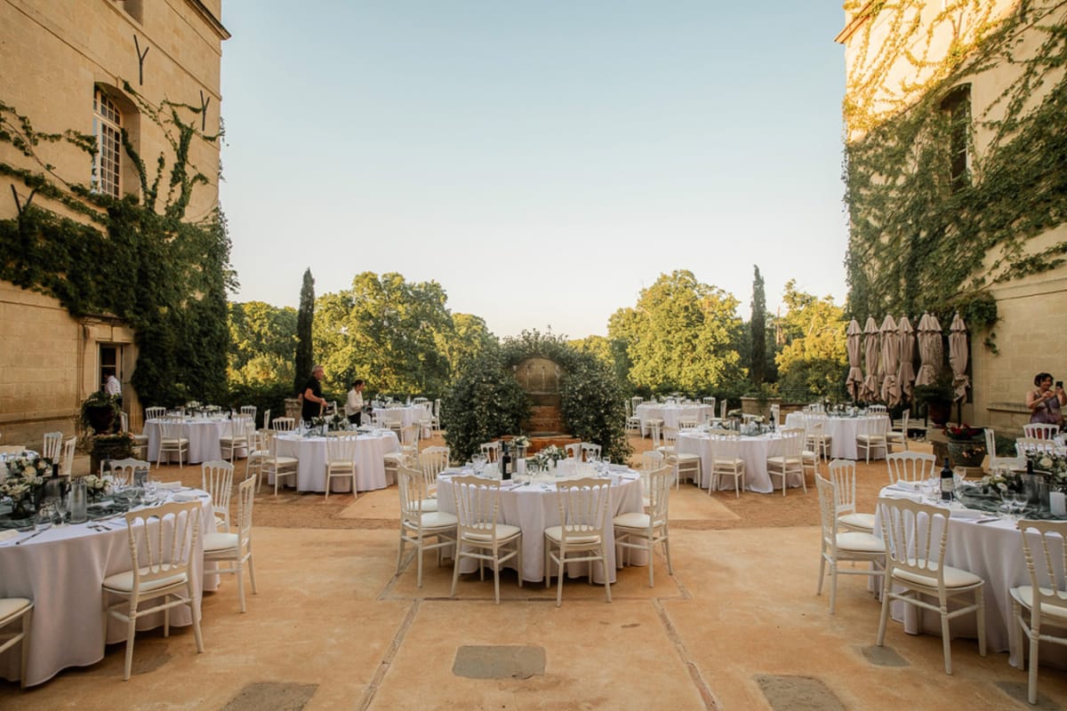 An outdoor wedding reception setup in the courtyard of a French château, photographed in a wide shot at golden hour. Approximately eight to ten round dining tables are arranged symmetrically across a stone-paved terrace, each dressed with white floor-length linen tablecloths and surrounded by white Chiavari chairs with cream cushions. Table centerpieces feature low arrangements of white and ivory flowers with greenery. A lush green floral arch frames a doorway at the far end of the courtyard, forming a natural focal point along the central aisle between the tables. Two staff members are visible making final preparations near the tables, and a guest photographs the scene from the right side. Folded taupe parasols are grouped to the right. The decor palette is white, ivory, and green, consistent with a classic, garden-style aesthetic. Potential venue feature image.