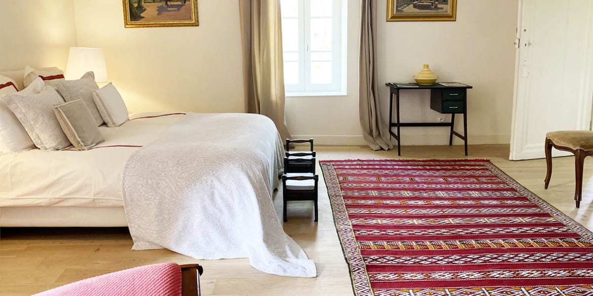Eclectic chateau bedroom with kilim rug, green writing desk, gold-framed paintings, and white linens